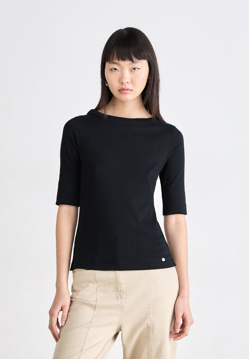 Svart kortermet topp med båthals, myk tekstur og tettsittende design. Har en liten logo ved kanten. Kombinert med lyse beige bukser.