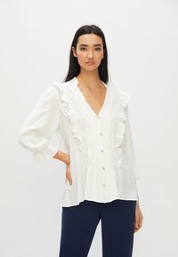 Blusa bianca con dettagli arricciati, scollo a V e chiusura con bottoni. Tessuto morbido, maniche lunghe con polsini arricciati e vestibilità rilassata.