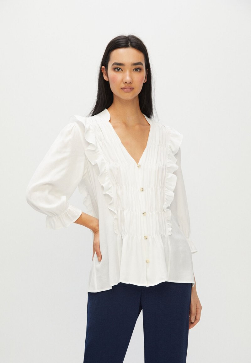 Blusa bianca con dettagli arricciati, scollo a V e chiusura con bottoni. Tessuto morbido, maniche lunghe con polsini arricciati e vestibilità rilassata.