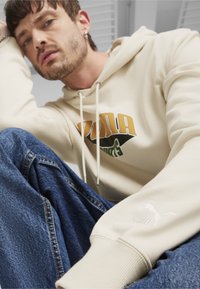 Felpa con cappuccio beige caratterizzata da una stampa grafica gialla e verde, abbinata a jeans di denim blu. Piccolo logo Puma bianco sulla manica.