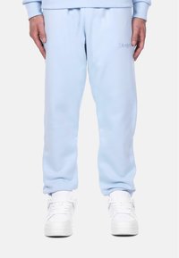 Light blue sweatpants in soft fabric with elastic cuffs, featuring an embroidered logo on the left thigh and paired with white sneakers.