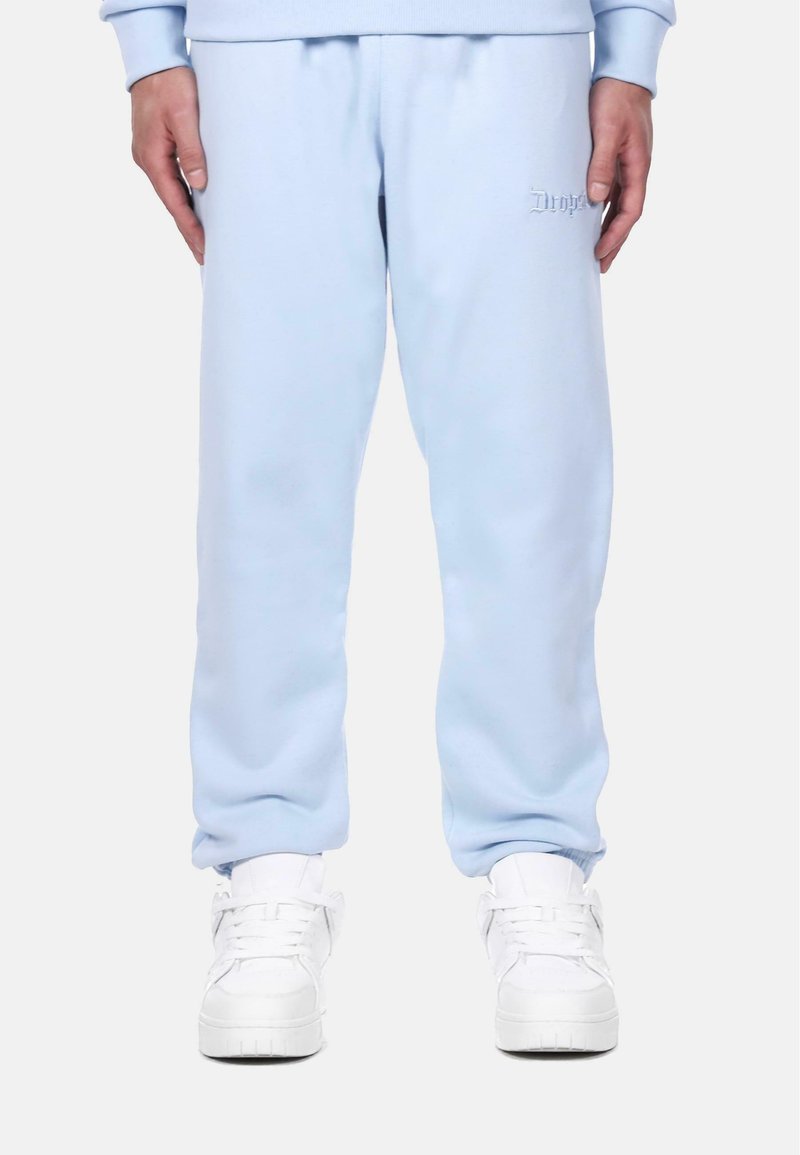 Helle blaue Sweatpants aus weichem Stoff mit elastischen Bündchen, ausgestattet mit einem gestickten Logo auf dem linken Oberschenkel und kombiniert mit weißen Sneakers.