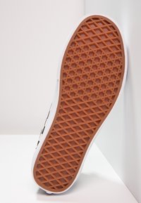 Semelle de basket blanche avec grip en caoutchouc marron à motif gaufré, inclinée contre un mur d’angle blanc et un sol.