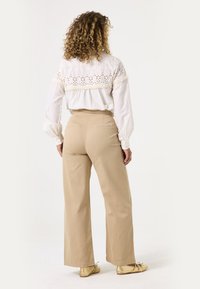 Weiße Bluse mit langen Ärmeln und gesticktem Einsatz, kombiniert mit beigen weit geschnittenen Hosen. Das Model steht mit dem Rücken zur Kamera und trägt metallisch goldene Ballerinas.