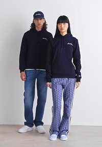 Mornarske majice sa kapuljačom u kombinaciji s plavim hlačama s uzorkom i bočnim prugama; muški model nosi traperice i usklađenu kapu, oba u bijelim tenisicama.