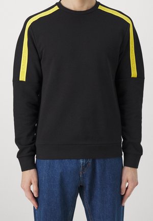 Schwarzes Sweatshirt mit langen Ärmeln und gelben Streifen auf den Schultern, mit dem Schriftzug "EA Emporio Armani", getragen zu blauen Jeans.