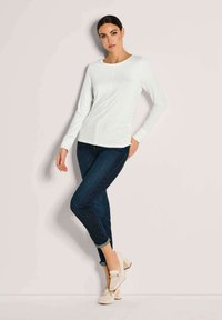 Frau, die an einer Wand posiert, trägt ein weißes langärmeliges Oberteil, dunkle hochgerollte Slim-Fit-Jeans und beigefarbene Blockabsatz-Schlüpfschuhe.
