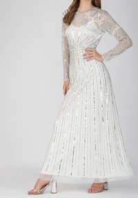 Abito maxi bianco con strisce di paillettes argentate, maniche lunghe trasparenti e dettagli ricamati sul corpetto. Sandali con tacco argentati completano il look.