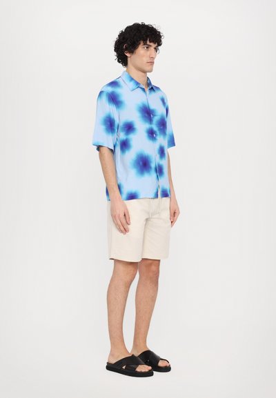 Cămașă cu mânecă scurtă, de culoare albastru deschis, cu modele florale albastre închise, combinată cu pantaloni scurți bej și sandale negre, stând pe un fundal alb.