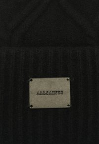 Tejido de lana negra con un patrón de ribetes texturizados, que presenta una etiqueta rectangular de cuero gris con el logotipo "ALLSAINTS" en relieve.