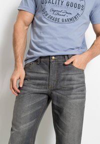 Jeans in denim grigio scuro con cuciture gialle a contrasto, dotati di una tasca frontale e chiusura con bottone metallico. Indossati con una t-shirt azzurra.