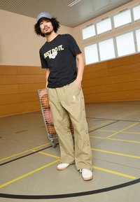 Svart T-skjorte med teksten "Just Do It", khaki tapered bukser med knappelukking, hvite sko med rosa såler, står ved siden av en vogn med basketballer.