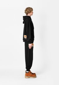 Conjunto de sudadera con capucha negra y pantalones de chándal, con bordado de osito de peluche en los bolsillos, hecho de una suave tela con un corte holgado. Botas marrones con cordones.