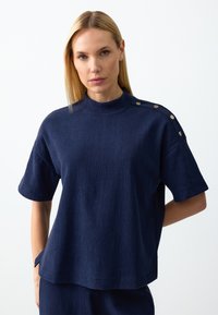 Blonde Frau, die ein navyfarbenes, strukturiertes Kurzarmoberteil mit hohem Kragen und metallenen Knopfdetails an den Schultern trägt, mit Händen hinter dem Rücken.