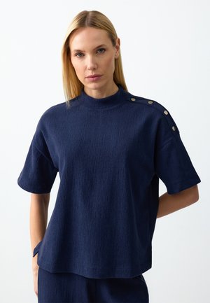 Blonde vrouw in een donkerblauwe, gestructureerde short-sleeve top met een hoge kraag en metalen knoopdetails op de schouder, handen achter de rug.