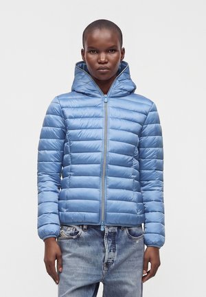 Person iført en lyseblå quiltet hættejakke med lynlås, kombineret med lyseblå vaskede jeans, stående mod en ensfarvet baggrund.