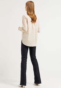 Camicia beige a maniche lunghe con motivo circolare, abbinata a jeans svasati scuri. La camicia presenta un orlo arrotondato e bottoni sul retro.