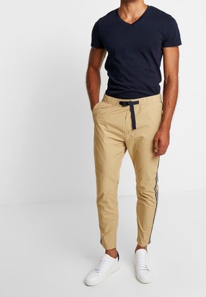 Granatowy t-shirt z dekoltem w serek, zestawiony z beżowymi zwężanymi spodniami z elastycznym pasem, granatowym sznurkiem i bocznymi paskami, uzupełnionymi białymi trampkami.