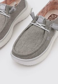 Zapatos slip-on de tela gris con un acabado texturizado, suela de goma y detalles en blanco en los cordones que contrastan. El interior presenta un forro con patrón.