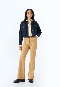 Chaqueta de denim azul oscuro con botones dorados, diseño acortado; combinada con pantalones de campana en tono beige claro y botas negras gruesas.