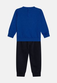 Camisa azul de manga larga con cuello redondo y cierre en la parte posterior con dos botones, combinada con pantalones de un azul marino oscuro que tienen puños elásticos en los tobillos.