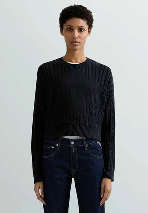 Donna che indossa un maglione nero a lavorazione a trecce con la scritta "REPLAY" e jeans blu scuro, in piedi davanti a uno sfondo semplice grigio chiaro.