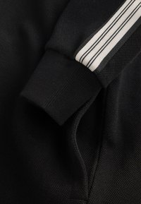 Tissu noir texturé avec poignet côtelé et détail de bandes blanches sur la manche d'un vêtement.