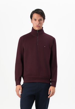 Man wearing a maroon quarter-zip pullover sweater and dark navy pants, standing against a plain white background.