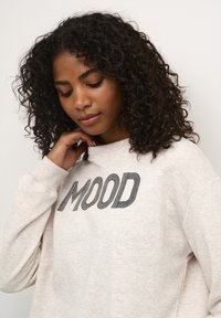 Helles beiges Sweatshirt mit strukturierter Oberfläche, versehen mit schwarzen gebänderten Buchstaben, die "MOOD" über die Brust spell. Lange Ärmel und runder Ausschnitt.