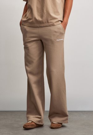 Personne portant un pantalon de survêtement beige ample, les mains dans les poches, et des chaussures marron à enfiler, debout sur un sol gris.