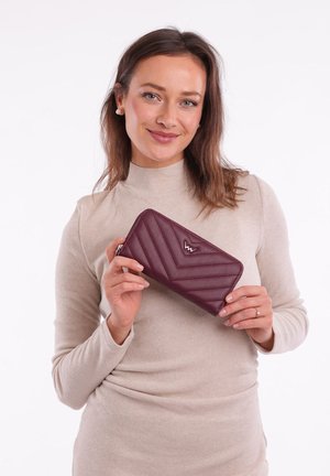 Burgundy leather wallet with quilted chevron pattern, silver logo accent, zip closure, held by a person wearing a beige sweater.