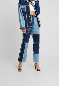 Kvinna klädd i en blå lapptäcktsjeansjacka och byxor med fransade kanter, vit kort topp och svarta klacksandaler, stående mot en grå bakgrund.