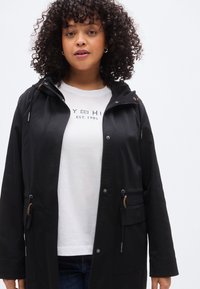 Μαύρο μπουφάν με κουκούλα, φερμουάρ στο μπροστινό μέρος, ρυθμιζόμενη μέση με κορδόνι και τσέπες με κόπιτσες. Διαθέτει ομαλή υφή και λεπτομέρειες σε ουδέτερους τόνους.