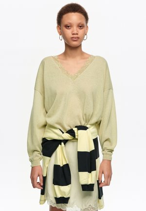 Persona con un suéter de cuello en V verde claro y una prenda a rayas negras y amarillas atada alrededor de la cintura, de pie frente a un fondo blanco liso.