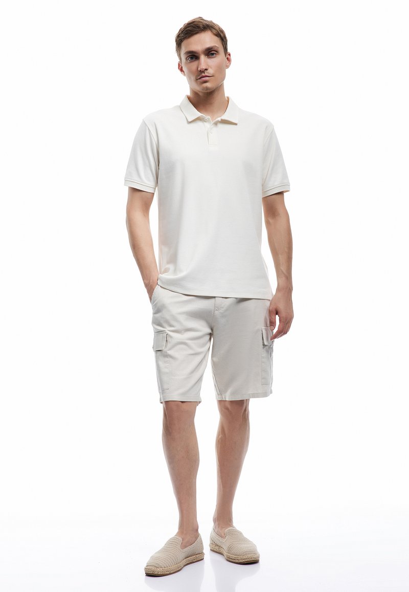 Camisa polo beige combinada con pantalones cortos de carga beige claro, ambos hechos de una tela suave. El conjunto presenta un diseño clásico y un estilo informal.