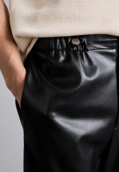 Schwarze Kunstlederhosen mit entspannter Passform, ausgestattet mit einem Kordelzugbund und subtilen Glanz. Beinhaltet ein Metallknopfdetail am Bund.