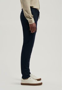 Persoon in een beige lange mouw shirt, marineblauwe fitted broek en witte sneakers met bruine zolen, zijwaarts staand op een effen achtergrond.