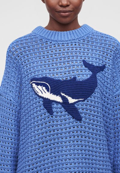 Personne portant un pull bleu tricoté avec un motif à maille ouverte et un grand motif de baleine bleu foncé et blanc sur la poitrine.