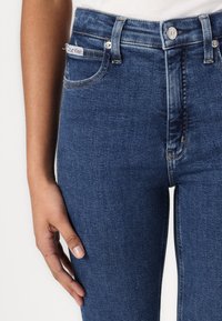 Tmavě modré džíny s úzkým střihem, s předním zapínáním na knoflík, pěti kapsami a malým logem CK na pásku.