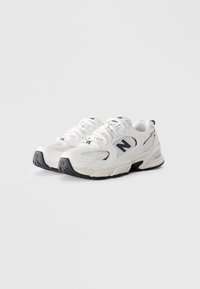 Une paire de baskets de sport blanches avec des semelles noires et le logo New Balance, inclinées pour montrer l'avant et les côtés sur un fond blanc uni.