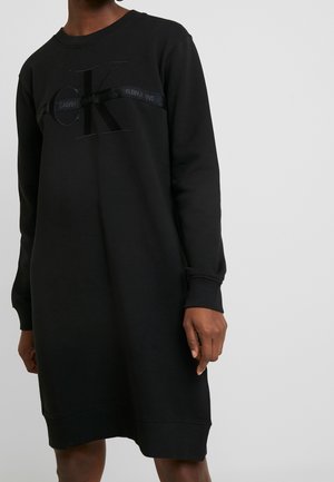 Osoba ubrana w czarną sukienkę dresową z długim rękawem, dużym logo CK i paskiem "Calvin Klein Jeans" na wysokości klatki piersiowej na jednolitym tle.