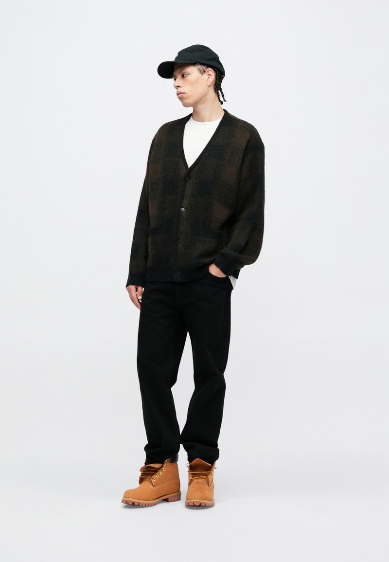 Black and brown plaid cardigan with a v-neck, button closure, two pockets, worn over a white shirt, paired with black pants and tan boots.