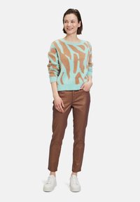 Mintgrüner Pullover mit braunem abstrakten Muster, kombiniert mit glänzenden, kupferfarbenen Hosen und weißen Sneakers. Weiches, strukturiertes Material überall.