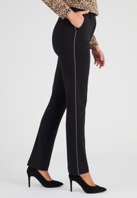 Pantalones negros ajustados con un corte slim, con una franja lateral en dorado que contrasta. Fabricados en un tejido suave, combinados con zapatos de tacón negros.