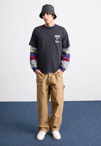 Tricou grafic negru, suprapus peste un tricou cu maneci lungi în dungi multicolore, asortat cu pantaloni cargo bej și sneakers albi, completat cu o pălărie tip bucket neagră.