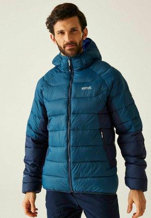 Mężczyzna w niebiesko-granatowej kurtce puchowej z kapturem, stojący na jasnym, jednolitym tle, patrzący w kamerę.