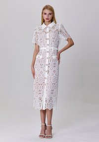 Robe en dentelle blanche avec des manches courtes, boutonnage devant et taille cintrée. Présente des motifs floraux, deux poches avant et un ourlet en dents de scie.