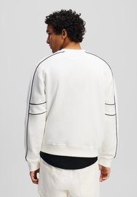 Sudadera blanca con detalles de piping negro a lo largo de las mangas y el cuello. Presenta un ajuste holgado y puños acanalados, llevada sobre una camisa negra.