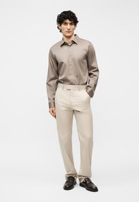 Νεαρός άνδρας στέκεται με το χέρι στην τσέπη φορώντας πουκάμισο ταφ με κουμπιά, μπεζ παντελόνι και σκούρα καφέ επίσημα παπούτσια σε λευκό φόντο.