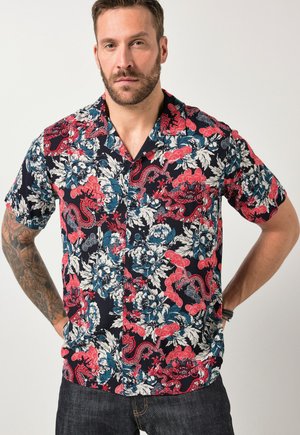 Mann mit Bart und Tätowierung, der ein kurzärmeliges Hemd mit roten, blauen und weißen Blumen- und Paisley-Mustern trägt, steht mit den Händen in die Hüften gestützt.